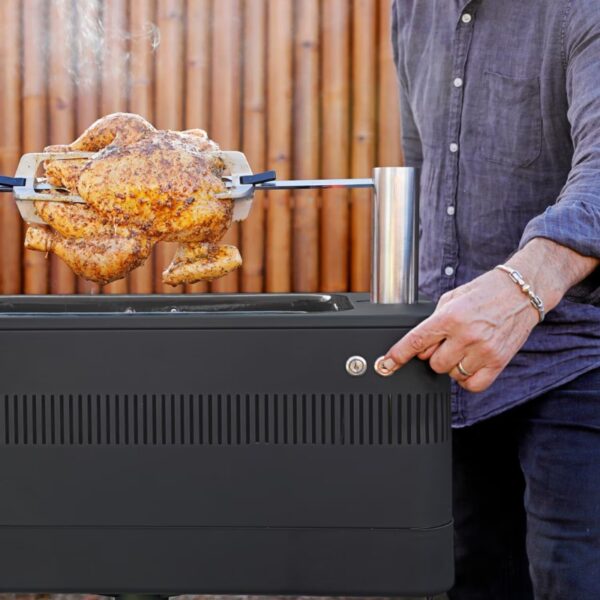 Man using a rotisserie grill to cook a seasoned chicken outdoors.