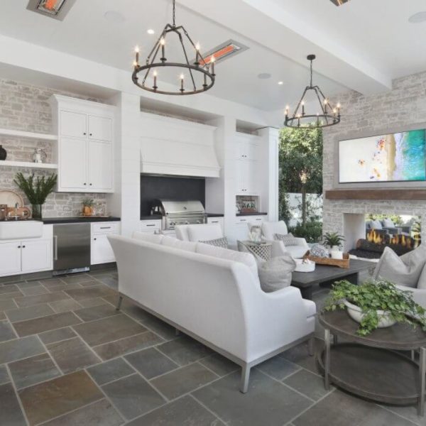Modern living room with a white sectional sofa, stone fireplace, and open kitchen featuring stainless steel appliances. Chandeliers provide ambient lighting, and large windows bring in natural light.
