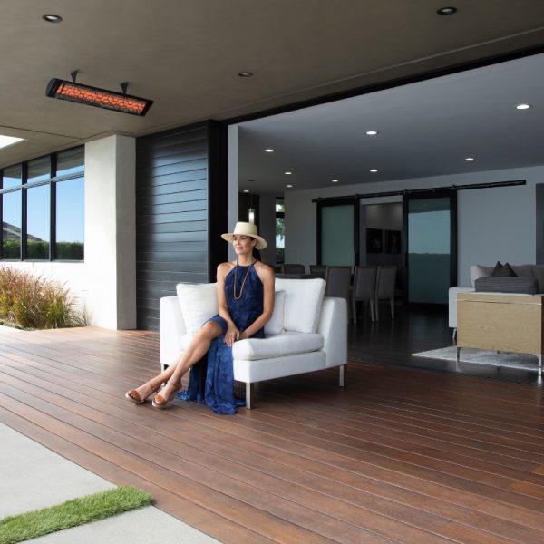 Woman in a blue dress and wide-brimmed hat relaxing on a white sofa on a modern patio, with an indoor dining area visible in the background.