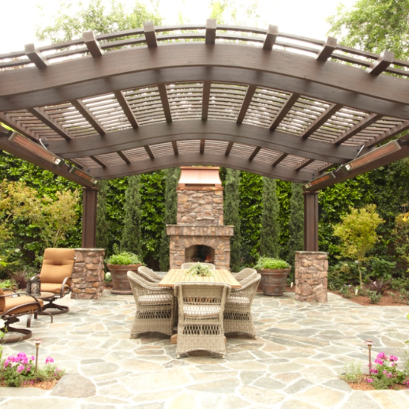 Outdoor patio featuring a wooden pergola, stone fireplace, and a dining table surrounded by wicker chairs, set in a lush garden.