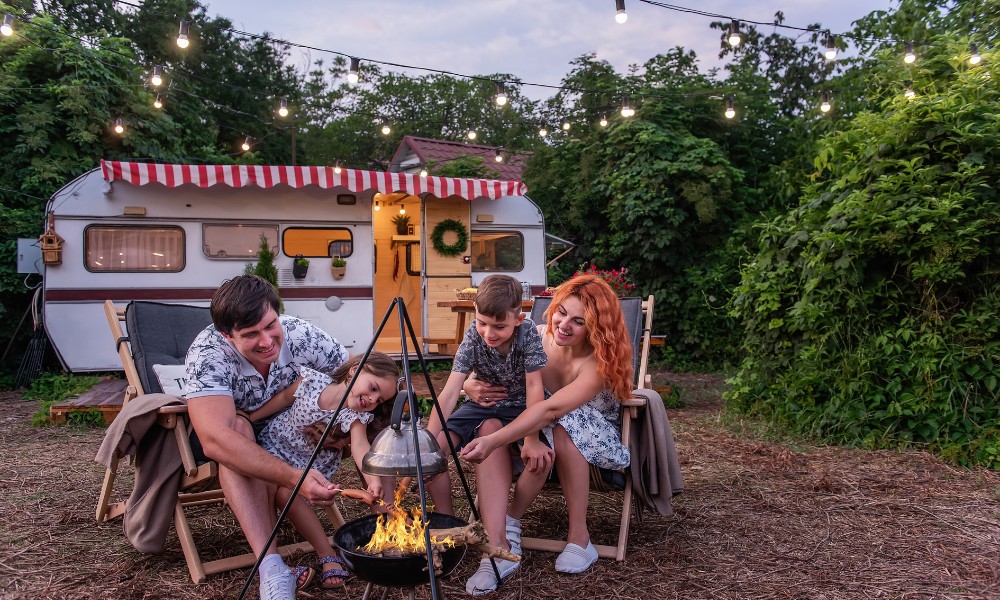 Family camping by a vintage caravan, enjoying a cozy evening around a campfire with string lights overhead.