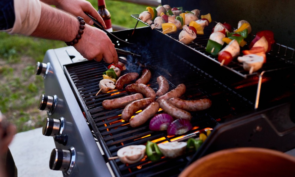 Person grilling sausages and colorful vegetable skewers on a barbecue, with smoke rising in an outdoor setting.