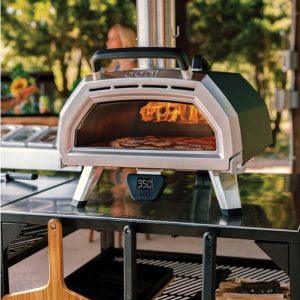 Ooni pizza oven with flames visible inside, displaying a pizza cooking at 950°F, placed on a stainless steel table in a backyard setting.