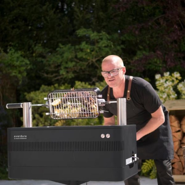 Man grilling food on an Everdure barbecue in a lush outdoor setting, showcasing the grill's rotisserie feature.