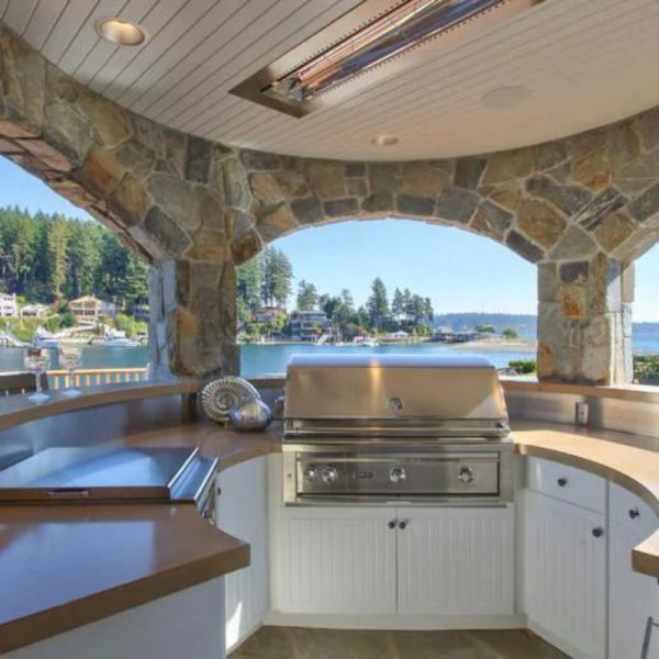 Outdoor kitchen with stone archway, featuring a stainless steel grill and scenic waterfront view. Ideal for entertaining and cooking in a beautiful setting.