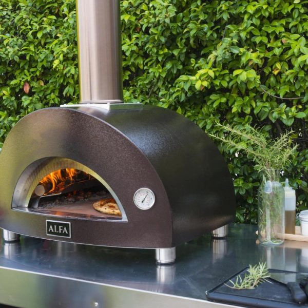 Outdoor pizza oven with a sleek design, featuring a visible fire and pizza inside, set against a lush green backdrop.