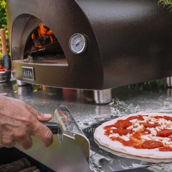 Hand using a metal pizza peel to transfer a pepperoni pizza topped with cheese and sauce to an Alfa pizza oven, with visible flames inside the oven and a thermometer on the front.