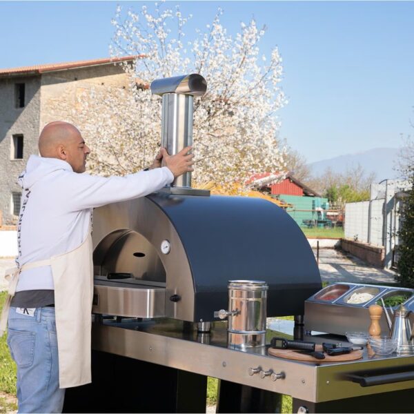 Person installing the chimney on a modern outdoor pizza oven in a garden setting, surrounded by blooming trees and kitchen prep tools.
