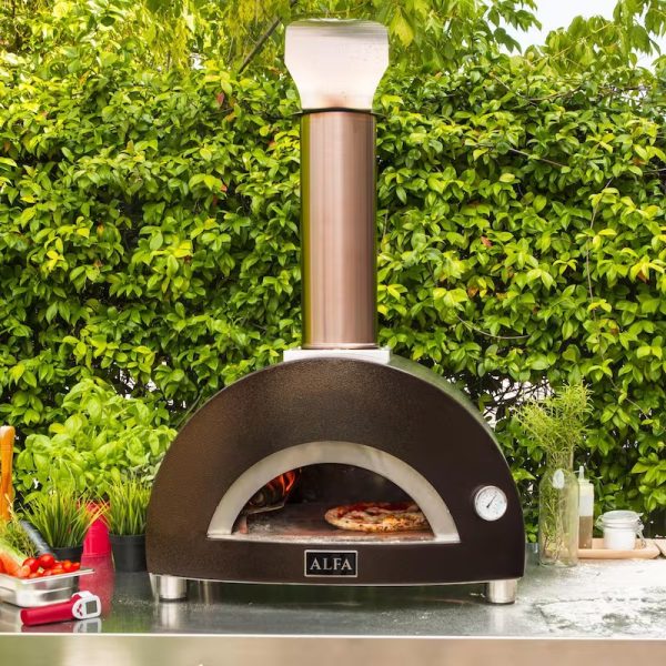 Outdoor pizza oven from Alfa with a visible flame and pizza inside, surrounded by greenery and cooking ingredients.