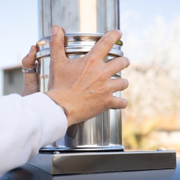 Person securing a stainless steel chimney cap on top of a chimney pipe outdoors.