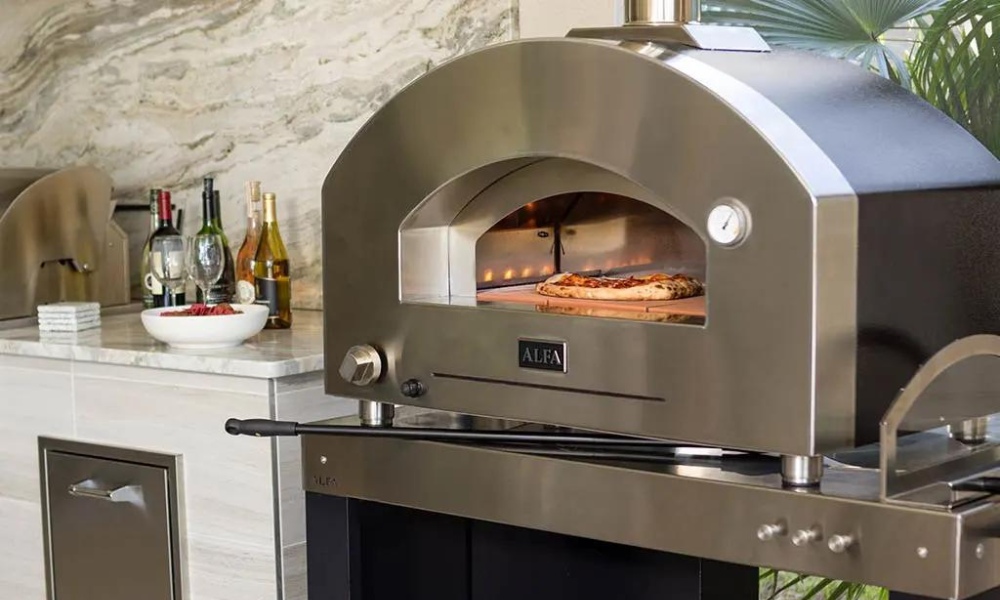 Stainless steel outdoor pizza oven with a pizza baking inside, set against a stylish kitchen backdrop featuring bottles of wine and a marble countertop.
