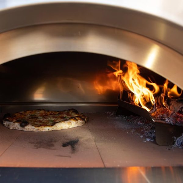 Wood-fired pizza cooking in a brick oven with flames and glowing embers.