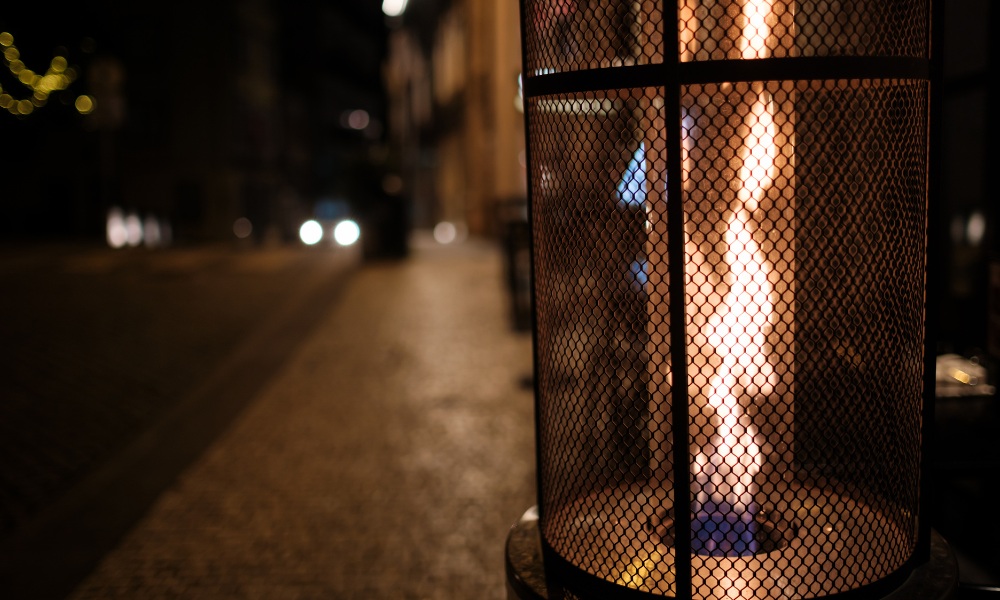 Outdoor flame heater illuminating a cobblestone street at night, with warm glow and blurred background lights.