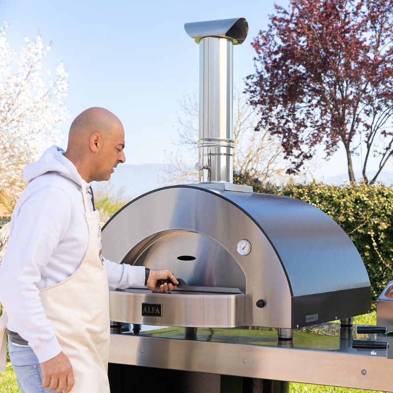 Man operating an Alfa outdoor pizza oven in a garden setting, showcasing its stainless steel design and chimney.