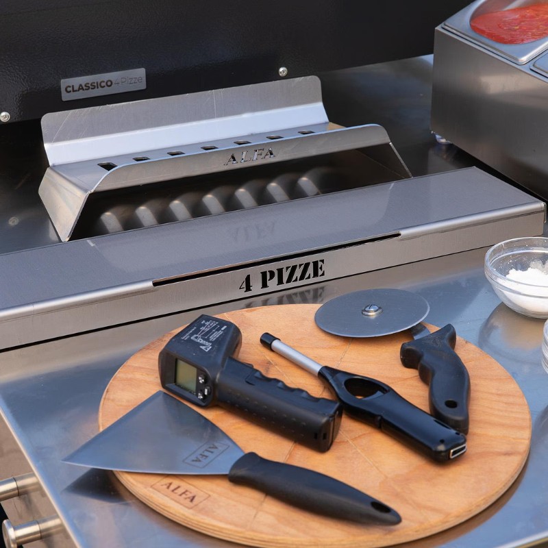 Stainless steel pizza oven with cooking tools, including a pizza cutter, thermometer, and spatula, on a wooden board.