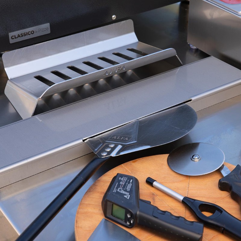 Stainless steel pizza oven tools including a pizza peel, cutter, and thermometer on a wooden board, with an Alfa pizza oven in the background.
