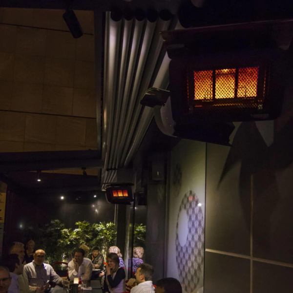 Interior view of a cozy restaurant with hanging infrared heaters, patrons engaged in conversation, and greenery in the background, creating a warm atmosphere.