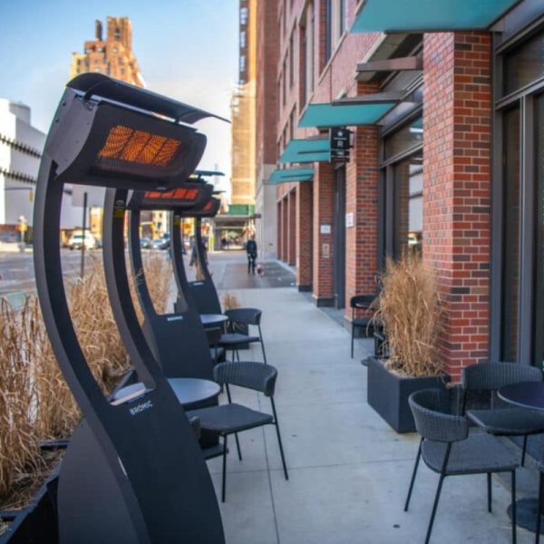 Outdoor seating area with infrared heaters along a city sidewalk, featuring modern black chairs and tables near a brick building.