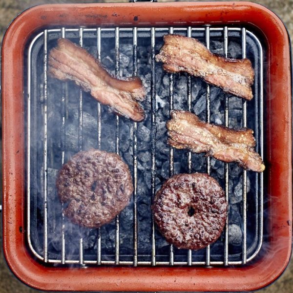 Grilling beef patties and bacon on a charcoal barbecue, with smoke rising from the hot coals.