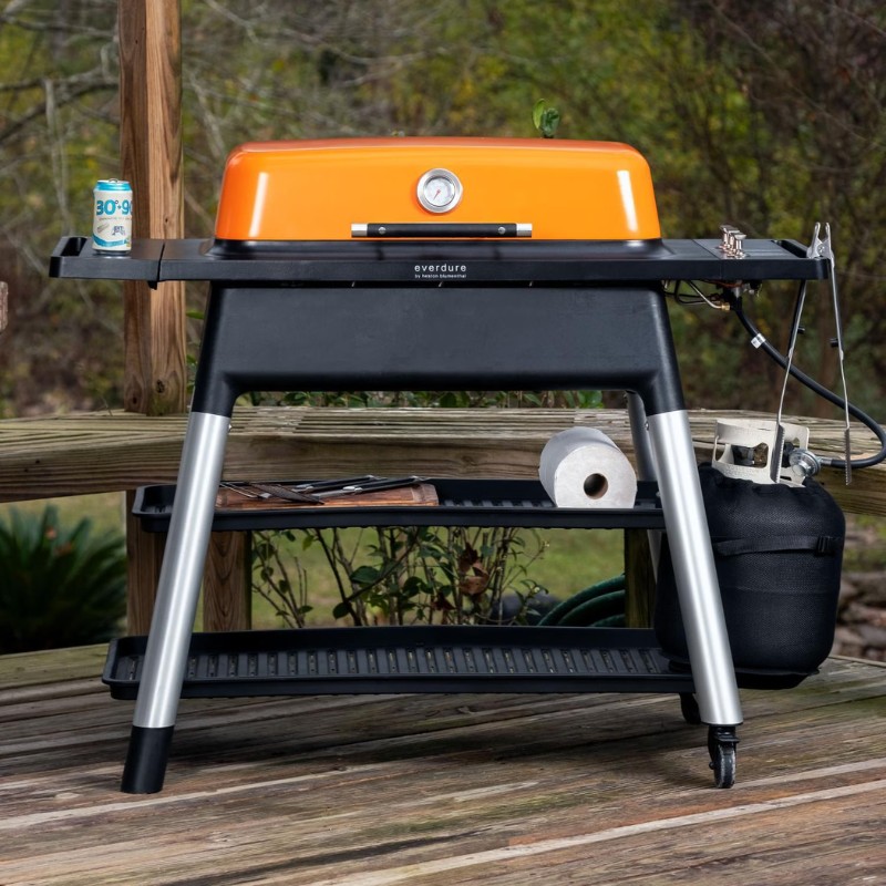 Portable gas grill with an orange lid, featuring a temperature gauge and storage shelves, set on a wooden deck.