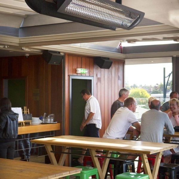 Interior of a lively bar with wooden walls, featuring patrons at a table and a staff member in a white shirt. Bright overhead heating lamps and colorful stools enhance the inviting atmosphere.