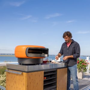 Man preparing food on a modern outdoor pizza oven with a scenic waterfront view.