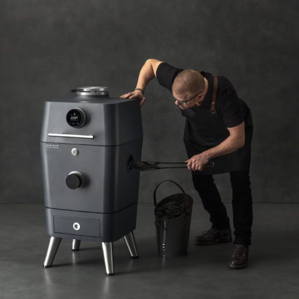 A chef adjusting the temperature on a modern charcoal smoker, demonstrating the cooking process with a bucket of charcoal nearby.
