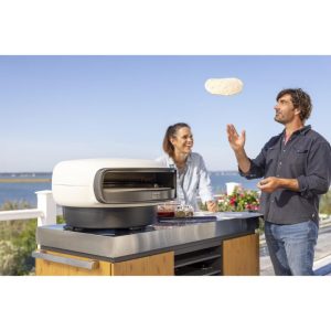 A man and woman enjoying outdoor cooking, with a pizza oven on a countertop, as the man tosses pizza dough in the air.