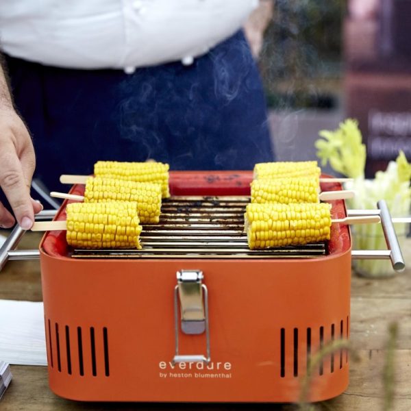 Grilled corn on the cob skewers cooking on an orange Everdure by Heston Blumenthal portable grill, with steam rising in a kitchen setting.
