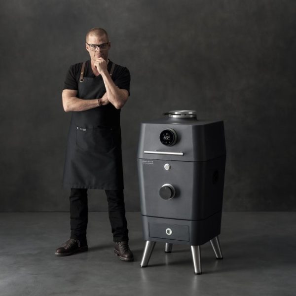 Chef in a black apron stands thoughtfully next to a modern gray cooking appliance with digital display, showcasing innovative cooking technology.