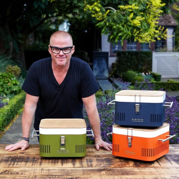 Chef showcasing Everdure outdoor cooking equipment in a garden setting, featuring colorful portable grills in green, navy, and orange.