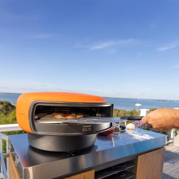 Outdoor pizza oven with vibrant orange top, being used to slide out a freshly baked pizza, set against a scenic waterfront backdrop.