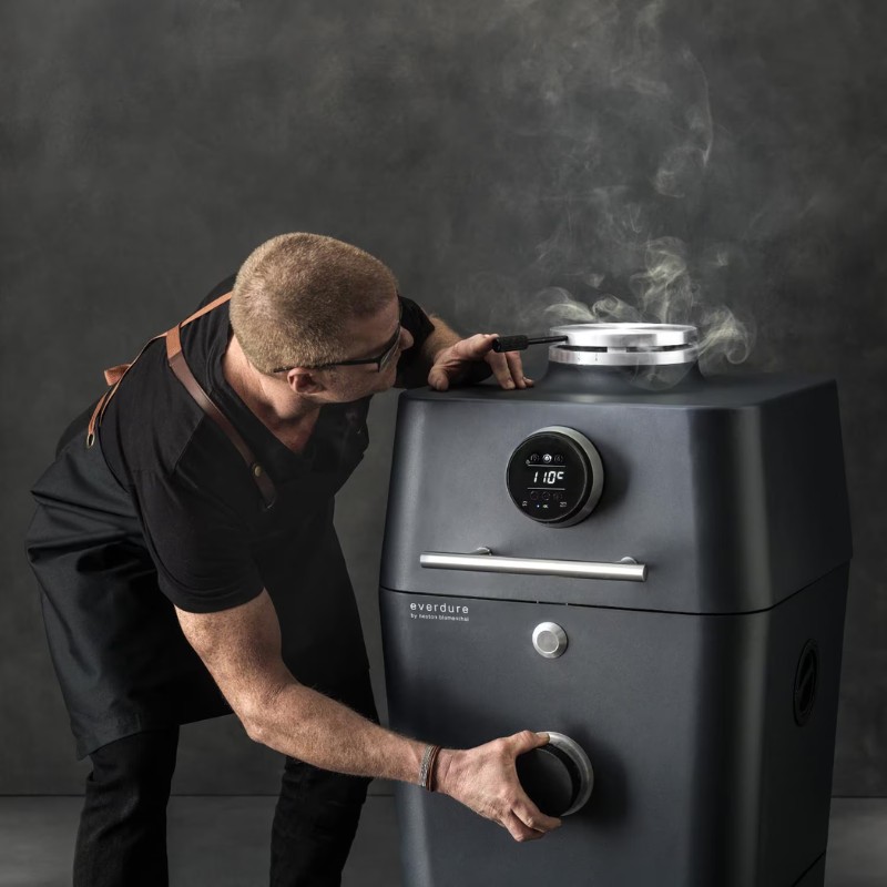 Man adjusting temperature settings on a modern Everdure outdoor smoker, with smoke rising from the top, showcasing advanced cooking technology.