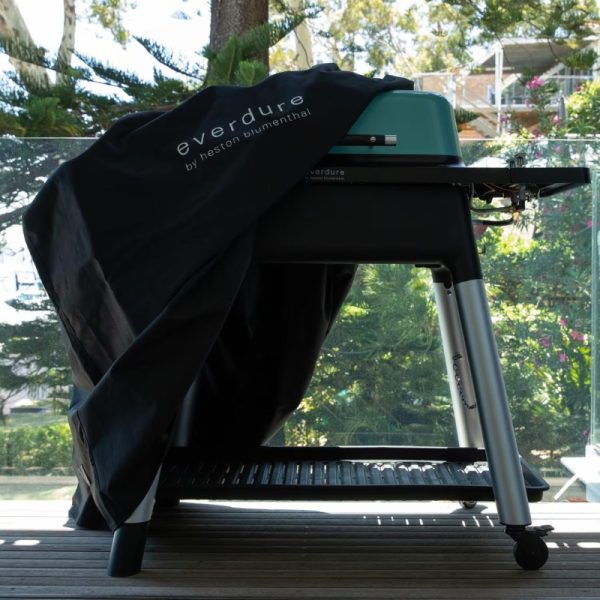 Everdure by Heston Blumenthal grill covered with a protective black cover, set against a backdrop of greenery and outdoor decking.