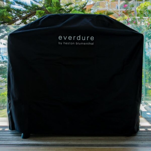 Black protective cover for an Everdure grill by Heston Blumenthal, placed on a wooden deck with a scenic background.
