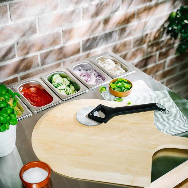 Wooden pizza peel with a pizza cutter on a kitchen countertop, featuring fresh ingredients like sliced zucchini, onions, and herbs in stainless steel containers against a rustic brick wall.