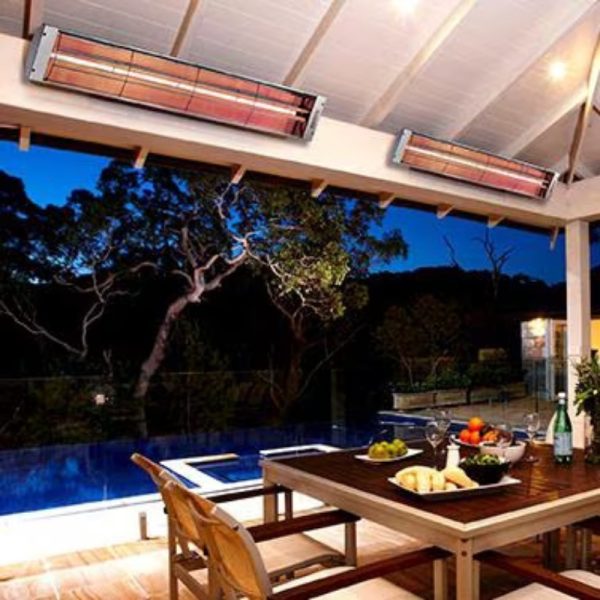 Outdoor dining area with a wooden table set, featuring fruit and drinks, illuminated by infrared heaters under a covered patio, with a swimming pool in the background at dusk.