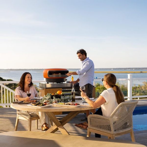 Outdoor gathering with friends enjoying a meal and wine, while a man grills on a modern barbecue by the water.