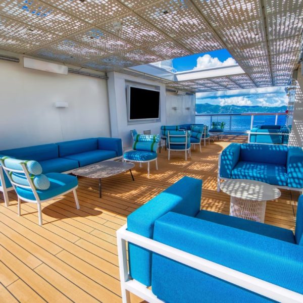 Modern outdoor lounge area with blue seating and wooden deck, featuring a shaded pergola and ocean view.