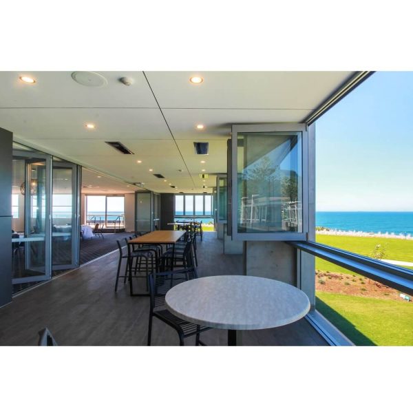 Modern interior of a beachfront cafe with large windows offering ocean views, featuring wooden flooring, tables, and chairs.