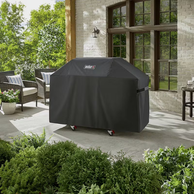 Weber grill covered with a black protective cover on a patio surrounded by greenery and outdoor furniture.