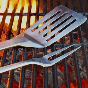 Grill tools, including a stainless steel spatula and fork, resting on a barbecue grill with glowing coals and flames.