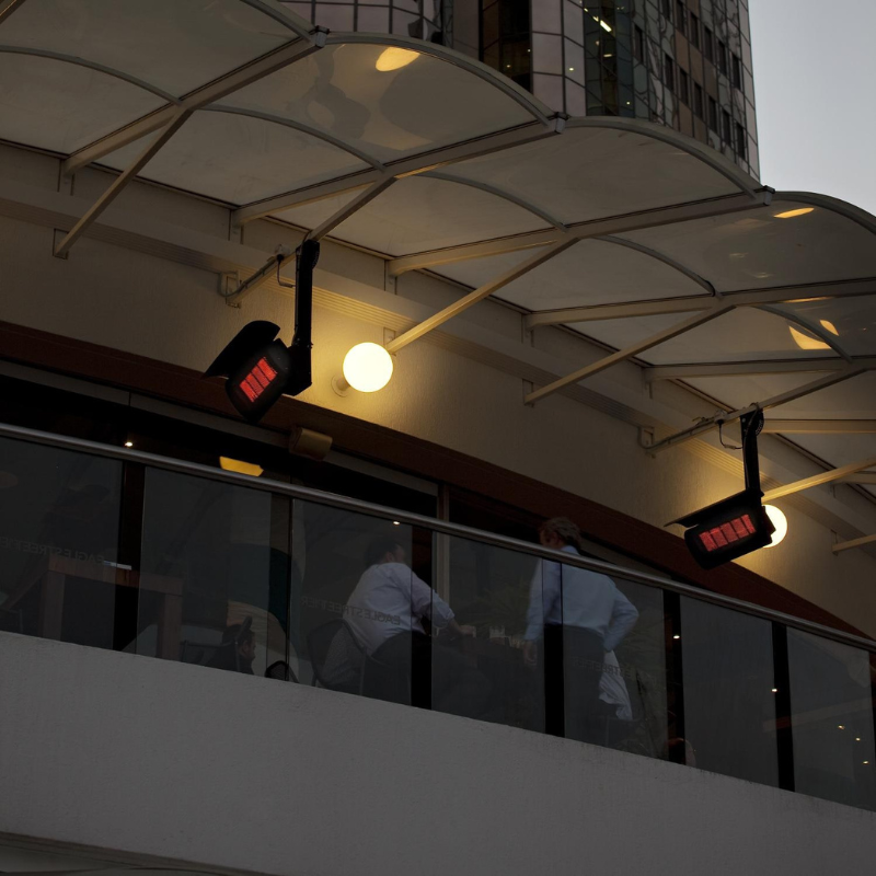 Exterior view of a modern building's balcony with overhead lights and red heating lamps, showcasing a social gathering.