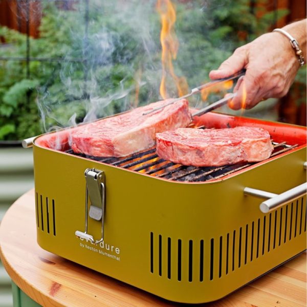 Hand holding tongs over a green portable grill with two steaks cooking and flames rising, surrounded by greenery.