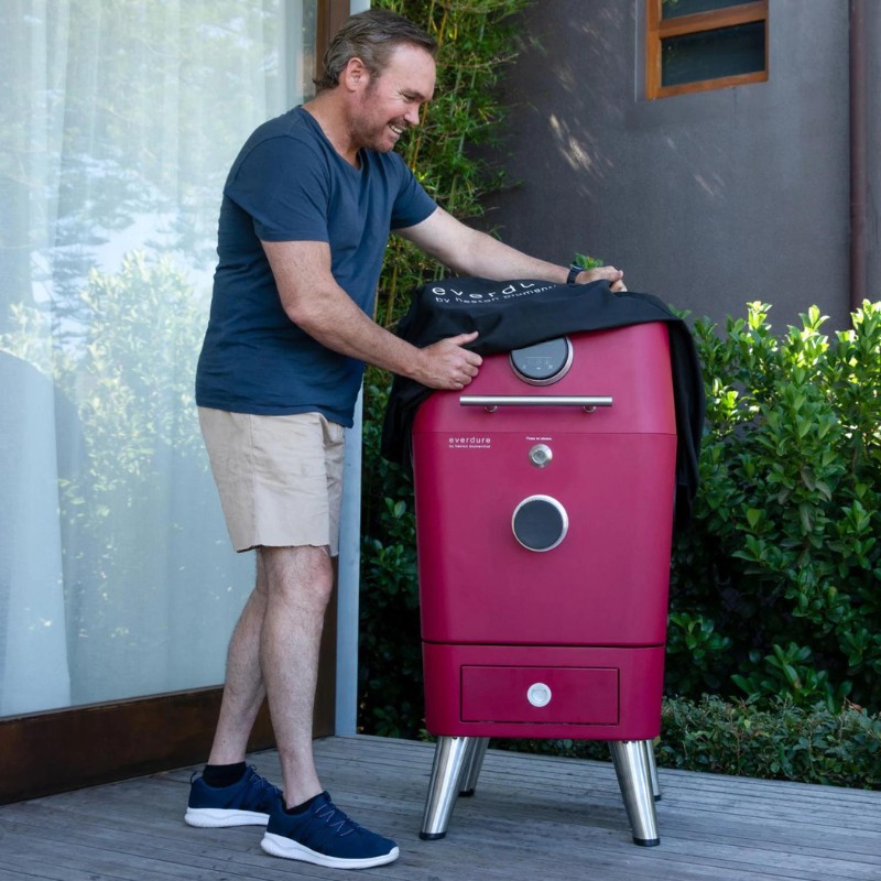 Man in casual attire preparing to use a red Everdure outdoor grill on a wooden deck surrounded by greenery.