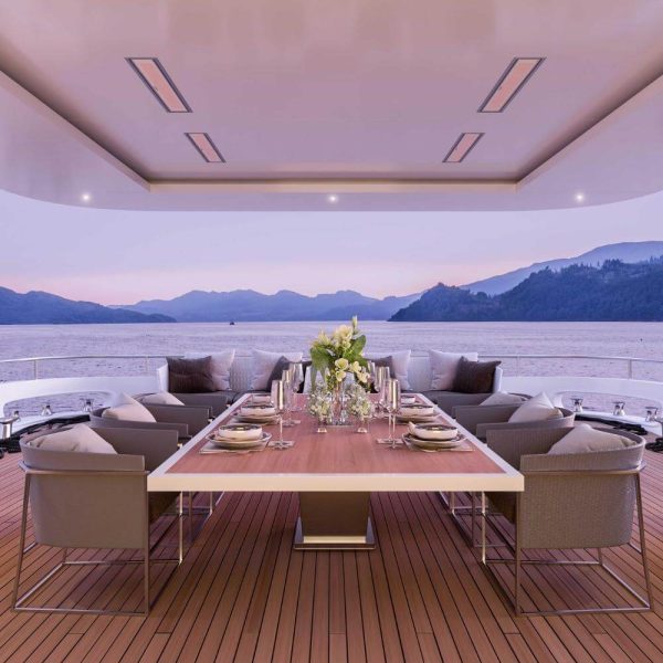 Elegant outdoor dining area on a yacht, featuring a large wooden table set with tableware and floral arrangements, overlooking a serene waterfront at sunset.