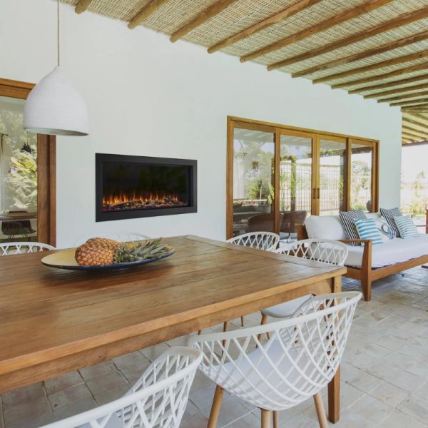 Modern outdoor dining area featuring a wooden table, white woven chairs, and a wall-mounted electric fireplace, with large glass doors leading to a scenic view.