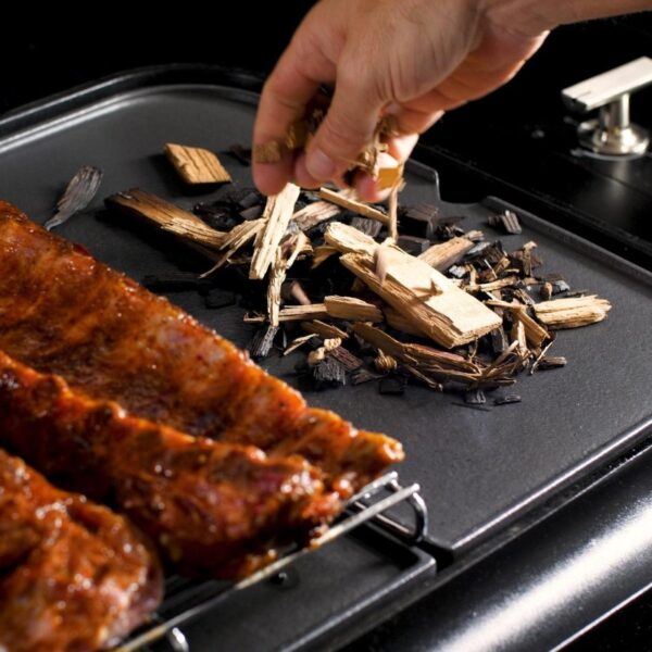 Hand adding wood chips for smoking on a grill with barbecue ribs in the foreground.