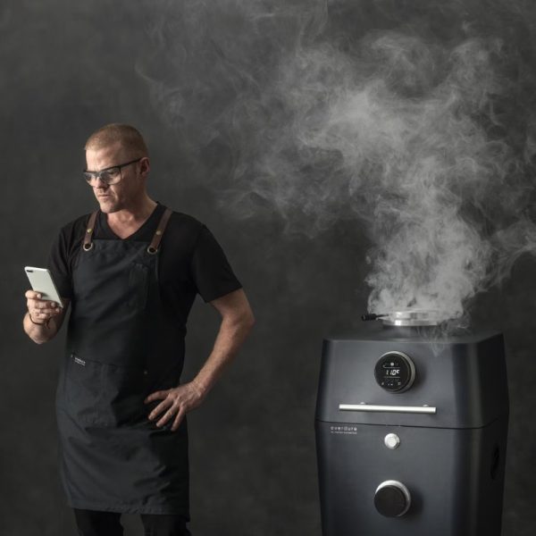 Chef in a black apron checks smartphone while standing next to a smoking Everdure cooker, emphasizing modern cooking techniques.