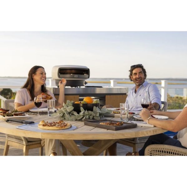 Outdoor dining scene with friends enjoying a meal, featuring a pizza and various dishes on a wooden table, with a grill in the background and a scenic view.